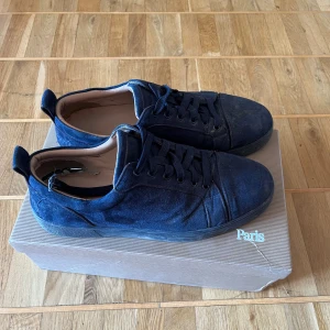 Mörkblå sneakers från Christian Louboutin - Säljer ett par mörkblå sneakers från Christian Louboutin med klassisk röd sula och svarta snören. Skorna har rund tå och är tillverkade i mocka. Perfekta för dig som vill ha en stilren och lyxig look.