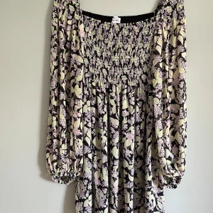 Blommig långärmad klänning från & Other Stories - Säljer en blommig långärmad klänning från & Other Stories med smockad överdel och elastiska ärmslut. En kortklänning som går till låren. Klänningen har svart bas med ljusa och lila blommor, och en luftig passform. Perfekt för vår och sommar!