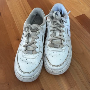 Vita Nike Air Force 1 sneakers - Klassiska vita Nike Air Force 1 sneakers med snörning och rund tå. Skorna har perforerade detaljer framtill och en platt sula. Perfekta för en stilren och sportig look.