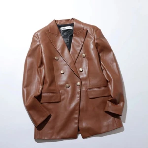 Brun skinnkavaj från Zara - Snygg brun kavaj i skinnimitation från Zara med dubbelknäppning och klassisk krage. Jackan har långa ärmar, två fickor framtill och guldiga knappar som detalj. Perfekt för en trendig och stilren look.