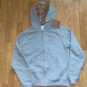 Grå hoodie från Burberry med dragkedja - Säljer en grå hoodie från Burberry med klassiskt rutigt foder i huvan och dragkedja framtill. Tröjan har fickor på sidorna, justerbar huva med snören och diskret Burberry-logga på bröstet.