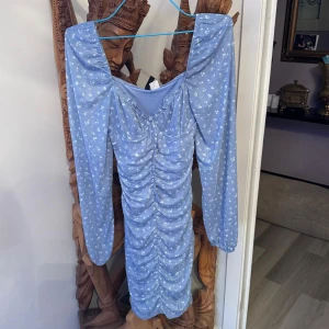 Blå blommig omlottklänning från Divided - Supersöt ljusblå kortklänning från Divided med små vita blommor, långärmad och med snygg rynkdetalj framtill. Offshoulder-modell som ger en trendig vibe. Perfekt för vår och sommar! endast använda 1 gång