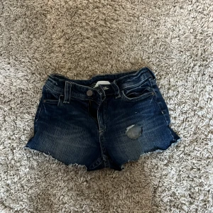 Mörkblå jeansshorts med slitning - Snygga mörkblå jeansshorts från Gina Tricot med råa kanter och en cool slitning på ena benet. Klassisk femficksmodell med knappar och bälteshällor. Perfekta för sommardagar när du vill ha en avslappnad och trendig look.