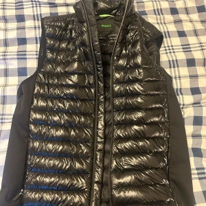 Svart dunväst från BOSS - Snygg svart dunväst från BOSS med glansig yta och quiltad design. Västen har hög krage och dragkedja framtill. Perfekt att slänga över en hoodie eller tröja för extra värme och stilpoäng.