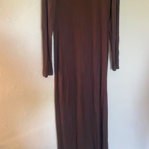 Brun långärmad maxiklänning AWARE - En stilren brun långklänning från AWARE med lång ärm och rak passform. Klänningen är enfärgad och har en enkel, minimalistisk look som passar perfekt för dig som gillar en clean och tidlös stil. Mjuk och skön i materialet, perfekt för alla årstider