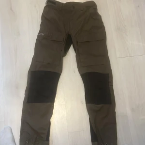 Bruna cargopants från Lundhags - Säljer ett par bruna cargopants från Lundhags med svarta förstärkningar över knäna. Byxorna har stora fickor på sidorna och broderad logga. Perfekta för dig som gillar outdoor och funktionella plagg. Och köpte in dom för 3200kr