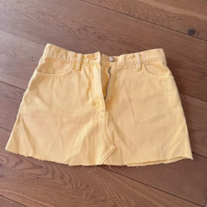 Gul jeanskjol med rå kant - Säljer en kort gul jeanskjol med rå, avklippt kant och klassiska fickor fram. Kjolen har knapp och dragkedja i midjan samt bälteshällor. Perfekt för dig som gillar en avslappnad och trendig stil.