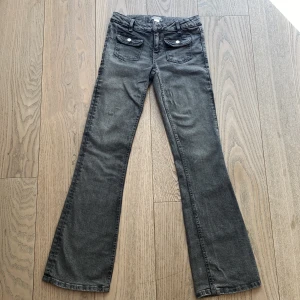 Grå Gina Tricot bootcut jeans med fickor fram - Säljer ett par grå jeans med bootcut passform. Byxorna har två fickor med lock och knapp framtill samt klassiska bakfickor. Jeansen har hällor för bälte och är i ett mjukt denimtyg. I stort sett oanvända.