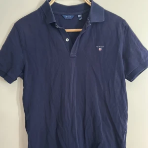 Mörkblå pikétröja från GANT - Säljer en klassisk mörkblå pikétröja från GANT med korta ärmar och krage. Tröjan har två knappar vid halsen och en liten broderad GANT-logga på bröstet. Perfekt för en stilren och avslappnad look. Passar även 168cm