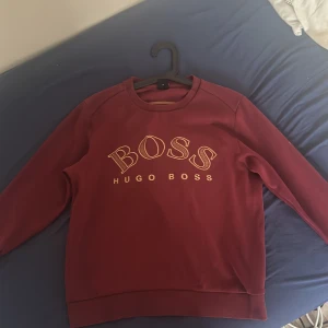 Vinröd sweatshirt från Hugo Boss - Snygg vinröd sweatshirt från Hugo Boss med stort BOSS-tryck i gult framtill. Tröjan har rund hals, långa ärmar och ribbade muddar. Perfekt för dig som gillar stilrena och trendiga plagg med premiumkänsla. Har en liten defekt på vänster arm.