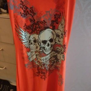 Orange ärmlös klänning med dödskalletryck - Cool orange kortklänning från Style med stort dödskalletryck och vingar framtill. Klänningen är ärmlös och har en snygg spetsdetalj i svart över ryggen. Perfekt för dig som gillar edgy och unika plagg.