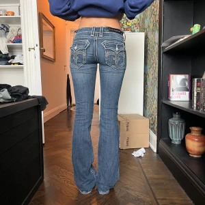 Rock Revival bootcut jeans blå - Snygga blå jeans från Rock Revival med bootcut passform och låg midja. Jeansen har tydliga kontrastsömmar, broderade bakfickor och detaljerade nitar. Tillverkade i klassiskt denim-material med coola detaljer som ger en unik look.