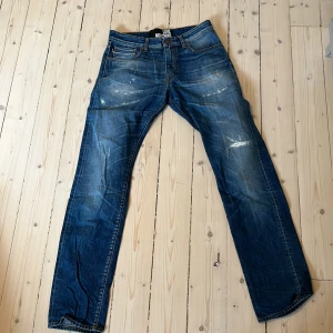 Blå jeans från Love Moschino - Snygga blå jeans från Love Moschino med coola slitningar och ett brunt peace-märke på bakfickan. Klassisk femficksmodell i bomull med raka ben och normal passform.