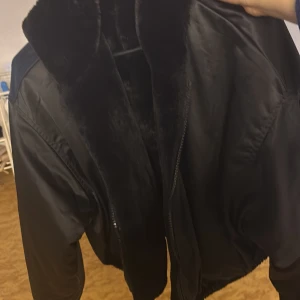 Svart pälsjacka med dragkedja - Säljer en svart pälsjacka med mjuk fuskpäls och dragkedja framtill. Jackan är kortare i modellen och har långa ärmar. Perfekt för dig som vill ha en stilren och mysig look under kyliga dagar.