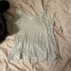 Ljusgrå linnetopp med spetsdetaljer - Supersöt ljusgrå linnetopp med tunna axelband och spets längs både ringning och nederkant. Dekorerad med en liten rosett framtill för extra gullig touch. Perfekt för lager-på-lager eller sommarens varma dagar.