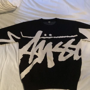 Svartvit stickad Stüssy tröja - Säljer en svart stickad tröja från Stüssy med stort vitt grafiskt logotryck över hela framsidan och ärmarna. Tröjan har rund halsringning och långa ärmar. Perfekt för dig som gillar streetwear och vill sticka ut med en fet logga. Aldrig använd, Passar flera olika stilar, går att göra mindre i torktumlaren, tvär skön och 100% ull