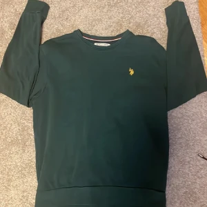 Grön polo tröja - Mörkgrön sweatshirt från Polo med klassisk rund hals och broderad gul logga på bröstet. Tröjan har långa ärmar och ribbade muddar vid ärmslut och nederkant. Perfekt för en chill och stilren look.