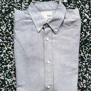 Grå skjorta från Ruffle & Bow L - Klassisk grå skjorta från Ruffle & Bow i storlek L. Skjortan har lång ärm, knappar hela vägen och en stilren krage med knappdetaljer. Perfekt för dig som gillar en enkel och clean look. Tillverkad i mjuk bomull som känns skön mot huden.