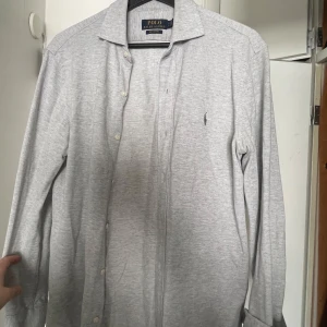 Ljusgrå skjorta från Polo Ralph Lauren -  Ljusgrå skjorta från Polo Ralph Lauren i riktigt nice material. Det som gör den riktigt riktigt nice är att den har betydligt bättre passform än helt nya då den är något ”kortare” i modellen. Även mer diskret broderad logga på bröstet. Och sen inga knappar vid kragen dvs ingen button up vilket är ovanligt för modellen. Sparsamt använd och i gott skick. 