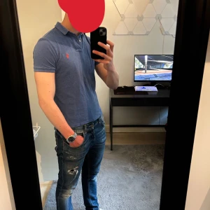 Ralph Lauren Piké - Säljer en klassisk blå pikétröja från Ralph Lauren med röd broderad logga på bröstet. Tröjan har korta ärmar, krage och knappar vid halsen. Perfekt för en stilren och avslappnad look.