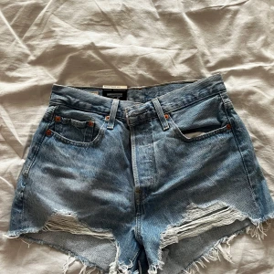 Levi's ljusblå jeansshorts med fransar - Säljer ett par Levi's ljusblå jeansshorts, modell Ribcage Straight Ankle, med råa fransade kanter och klassiska fem fickor. Hög midja och avslappnad passform, perfekt för sommardagar. Ikonisk Levi's-lapp bak och röd etikett på fickan.
