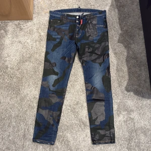 Camouflage jeans från Dsquared2 - Storlek 50 som motsvarar 34-36. Säljer ett par snygga jeans från Dsquared2 med blå och grön camouflage-mönster. Byxorna har klassisk femficksdesign, knappgylf och raka ben. Märkespatch i svart läder baktill i midjan. Perfekta för dig som vill sticka ut med din stil.