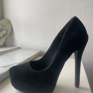 Svarta pumps med platå och hög klack - Säljer ett par svarta pumps med rund tå, platåsula och riktigt hög klack. Skorna är i mockaliknande material och har en stilren, klassisk design som passar perfekt till festliga tillfällen.