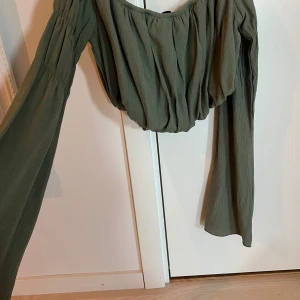 Khakigrön blus med vida ärmar - Säljer en khakigrön blus från NA-KD med vida, långa ärmar och rynkade detaljer. Toppen har en lös passform och är tillverkad i ett lätt, strukturerat tyg. Perfekt för dig som gillar trendiga och bekväma plagg.