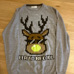 Grå stickad jultröja med renmotiv från H&M - Rolig grå stickad tröja från H&M med ett stort pixel-art renmotiv med solglasögon och texten 'DEER TO BE COOL' framtill. Perfekt för julens alla tillfällen. Långa ärmar och rund hals.