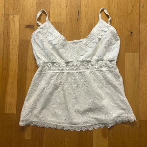 Vit sommar top - Säljer en söt vit linnetopp med broderade blommor och spetsdetaljer längs halsringningen och nederkanten. Toppen har smala axelband och en v-ringning framtill. Perfekt till sommaren! Den har en liten fläck, men den går troligen att tvätta bort (bild 4)