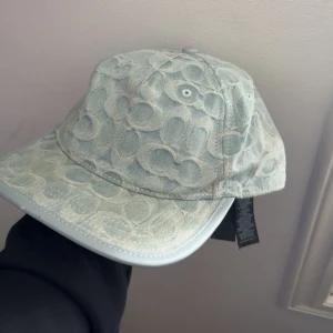 Coach Keps - Snygg ljusblå keps från Coach med mönster av Coach-loggor. Justerbart läderband baktill med metallspänne. Perfekt accessoar för en trendig look. 