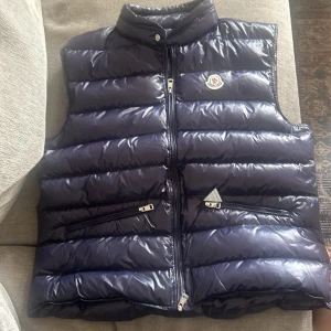 Blå dunväst från Moncler - Säljer en mörkblå, glansig dunväst från Moncler med ståkrage och dragkedja framtill. Västen har två snedställda fickor med dragkedja och Moncler-logga på bröstet. Perfekt för lager-på-lager under kyliga dagar.