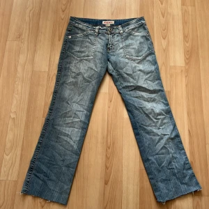 Blå bootcut jeans med rå kant - Säljer ett par klassiska blå jeans med bootcut passform och rå, ofållad kant nertill. Jeansen har fem fickor, dragkedja och knapp framtill samt ljusa slitningar för en avslappnad look. 