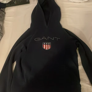 Mörkblå hoodie från GANT - Säljer en mörkblå hoodie från GANT med broderad logga och sköld på bröstet. Klassisk huva och långa ärmar, perfekt för en avslappnad stil. Mjuk insida och ribbade muddar vid ärmslut och nederkant.