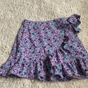 Blommig kjol med volang från FB Sister - Superfin blommig kjol från FB Sister i storlek S. Kjolen har volangdetaljer längs nederkanten och knytband i midjan. Färgerna går i lila, turkos, blått och grönt. Perfekt för dig som gillar färgglada och lekfulla plagg! Har även en dragkedja på sidan