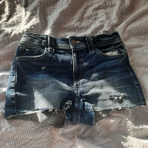Mörkblå jeansshorts med slitningar - Snygga mörkblå jeansshorts med råa, fransiga kanter och slitna detaljer framtill. Klassisk femficksmodell med knapp och dragkedja. Perfekta för sommardagar när du vill ha en avslappnad och cool stil. klippta hemma