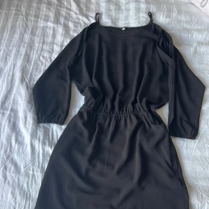 Svart kort klänning BY FE - Svart kort klänning från BY FE med trekvartsärm och tunna axelband. Klänningen har elastisk midja och en snygg cut-out detalj vid axlarna. Perfekt för dig som gillar stilren och enkel design med en twist.