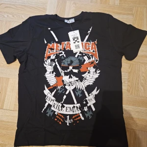 Svart Metallica x Off White t-shirt - Svart t-shirt från Off White i samarbete med Metallica. Stor tryckt grafik framtill med dödskalle, vingar och orange Metallica-logga. Coolt, edgy tryck och klassisk passform. Perfekt för dig som gillar streetwear och rock.