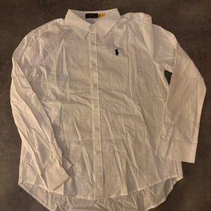 Vit linne skjorta från Polo Ralph Lauren - Klassisk vit skjorta från Polo Ralph Lauren med blå broderad logga på bröstet. Skjortan har lång ärm, knappar framtill och är tillverkad i ett tunt bomullstyg. Perfekt för en clean och stilren look. Linne skjorta 