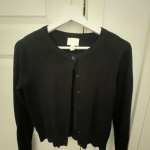 Svart kofta från H&M med knappar - En svart kofta från H&M med rund halsringning och knäppning framtill. Koftan har långa ärmar och en något croppad passform med vågig nederkant. Perfekt att slänga över en topp för en enkel och stilren look.