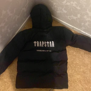 Svart pufferjacka från Trapstar - Säljer en svart pufferjacka från Trapstar med huva och dragkedja. Jackan har logga på bröstet och stor Trapstar-tryck på ryggen. Perfekt för kalla dagar när du vill hålla stilen.