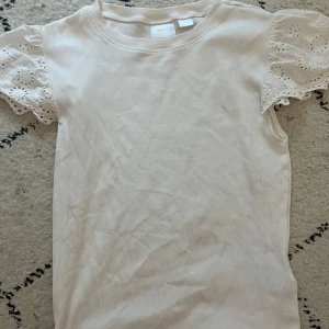 Vit ribbad topp med spetsärmar från Gina Tricot - Supersöt vit topp från Gina Tricot i ribbat material med korta ärmar i spets och broderade detaljer. Perfekt för en fräsch och feminin look. Rund halsringning och mjuk bomullskänsla.