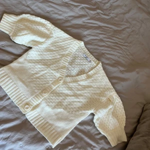 Vit stickad kofta från NA-KD - Säljer en söt vit stickad kofta från NA-KD med korta puffärmar och v-ringning. Koftan har ett fint mönster och knappar framtill. Perfekt för lager-på-lager-stil eller som ett lättare plagg under svalare dagar.