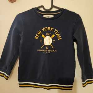 Mörkblå tröja från Hampton Republic - Säljer en mörkblå tröja från Hampton Republic med tryck 'New York Team' och två korsade basebollträn. Tröjan har långa ärmar och ribbade muddar med gula och vita ränder vid ärmslut och nederkant. Perfekt för en sportig look!