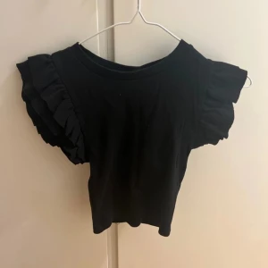 Svart topp med volangärmar - Säljer en svart topp med volangdetaljer på ärmarna. Toppen har en enkel design och är perfekt för en stilren look. Passar bra till både jeans och kjol.