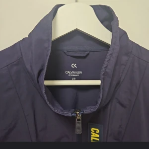 Flerfärgad vindjacka från Calvin Klein - Säljer en snygg flerfärgad vindjacka från Calvin Klein. Jackan har en dragkedja framtill och är långärmad. Den är i en kombination av mörkblått, gult och grått med ett stilrent märke på bröstet. Perfekt för en sportig look!