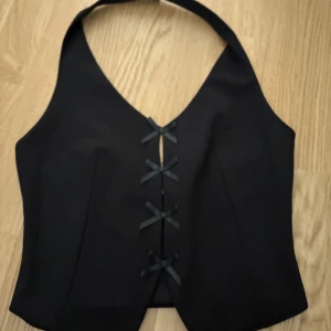 Svart korsettväst från Gina Tricot - Snygg svart korsettväst från Gina Tricot med knytning framtill. Perfekt för att ge en stilren touch till din outfit. Västen har en elegant design med en halterneck-stil. Använd en gång 