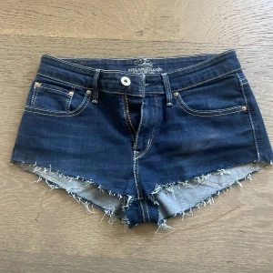 Mörkblå jeansshorts från Levi's - Snygga Lågmidjade  mörkblå jeansshorts från Levi's med fransiga kanter och klassisk femficksdesign. Perfekta för en avslappnad stil. De har en knapp och dragkedja framtill.