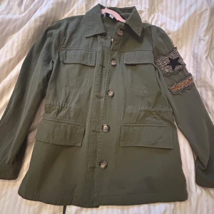 Olivgrön jacka från CIMINY - Snygg olivgrön jacka med kappar, från märket CIMINY Paris i storlek Xs. Aldrig använd. Fina detaljer på ena armen, 4 fickor på jackan 