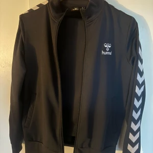 Svart träningsjacka från Hummel och Adidas - Två svarta träningsjackor, en från Hummel med vita chevronmönster på ärmarna och en från Adidas med klassiska ränder. Båda har dragkedja och långärmade design. Perfekta för sport och vardag. S-M två för 400kr 300kr/st